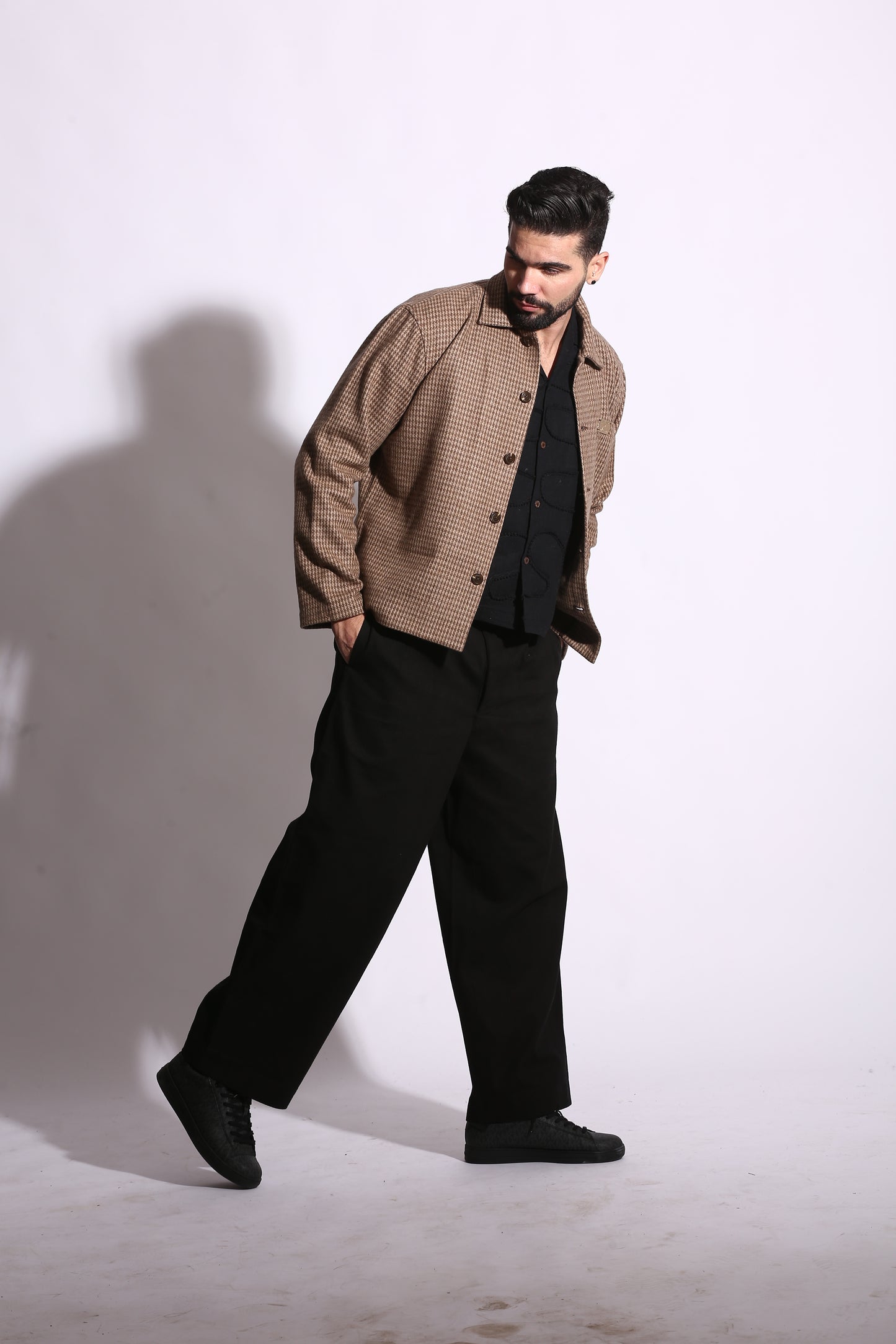 Man wearing a brown jacket and black pants standing against a white wall.
