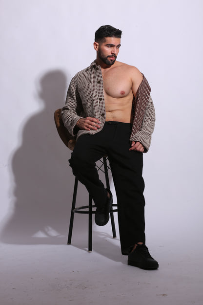 Man sitting on a chair without a shirt, wearing a patterned jacket and black pants.