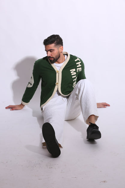 Person wearing a green sweater with text and white pants on a white floor.