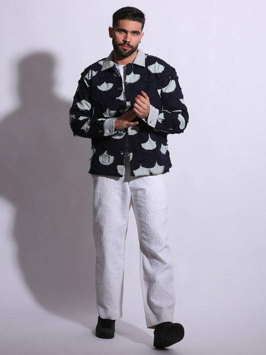 Man wearing a dark jacket with white patterns and light pants on a plain background