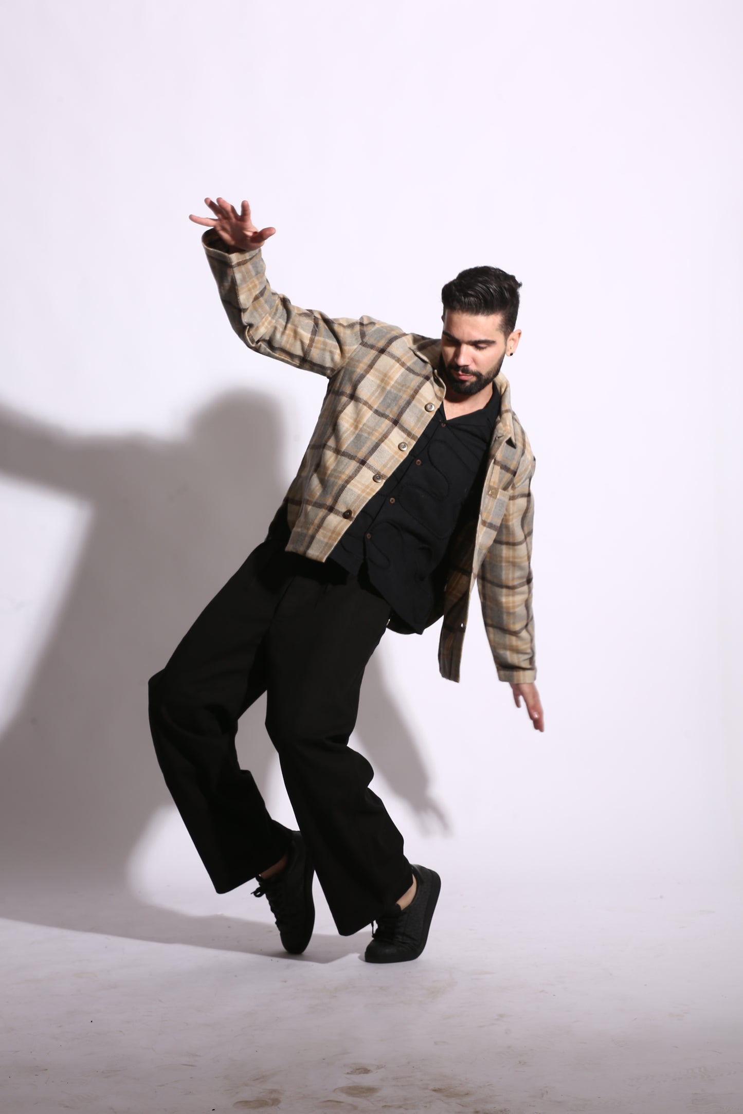 Person in plaid shirt and black pants standing on a white floor with a shadow cast on the wall.