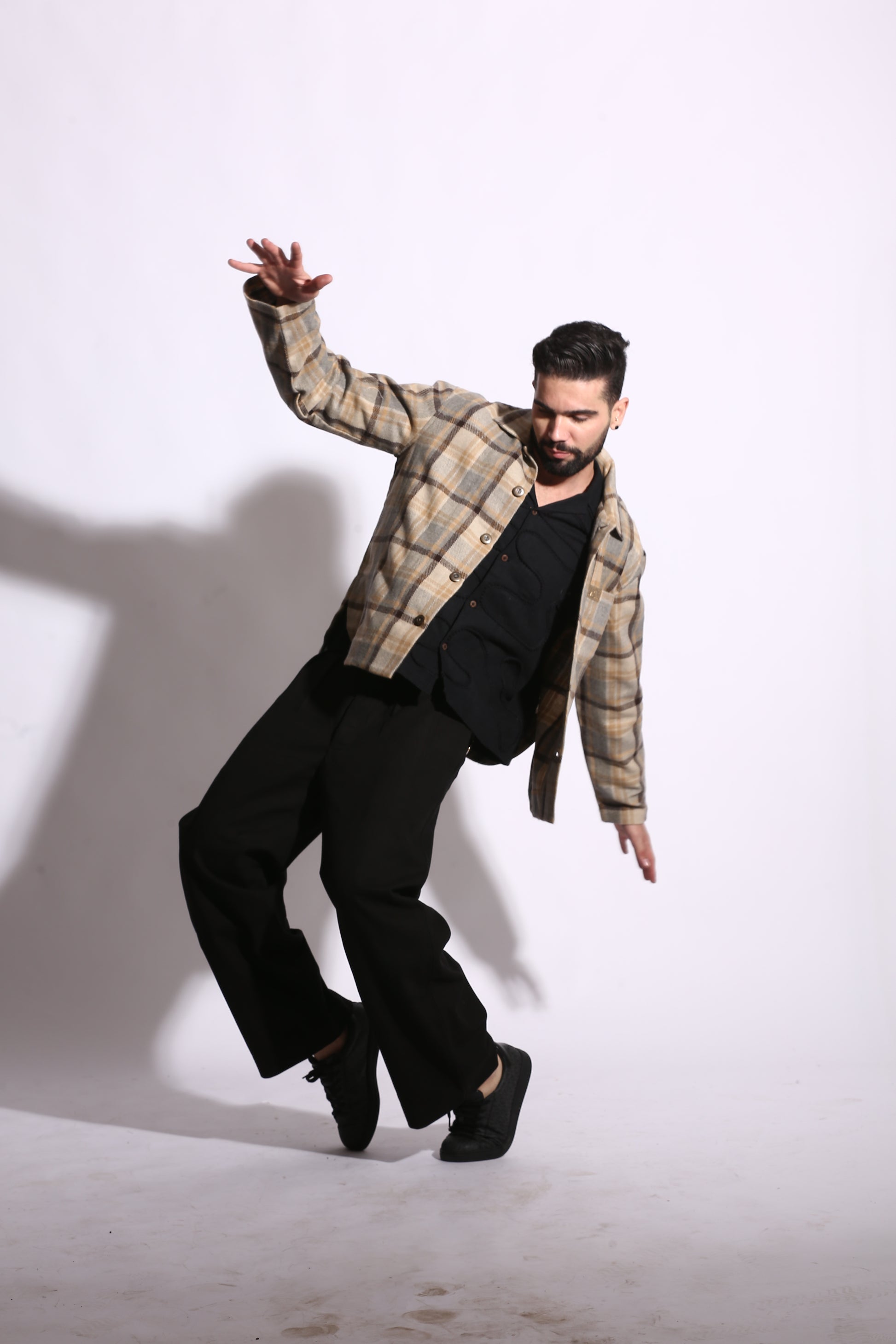 Person in plaid shirt and black pants standing on a white floor with a shadow cast on the wall.