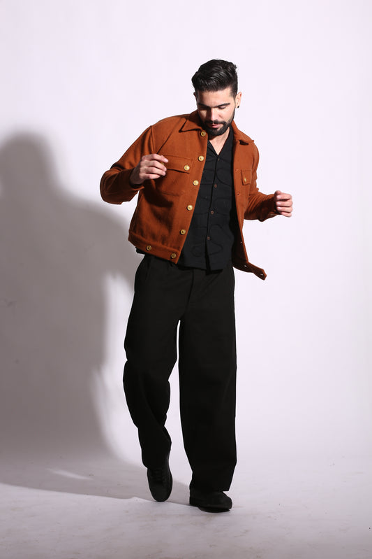 Person wearing a brown jacket and black pants standing on a white floor with a shadow cast on the wall.