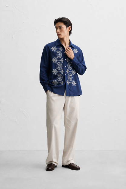 A man wearing a navy blue long-sleeve button-up shirt with white floral embroidery on the front, paired with off-white trousers and dark brown loafers, standing against a light gray textured wall.