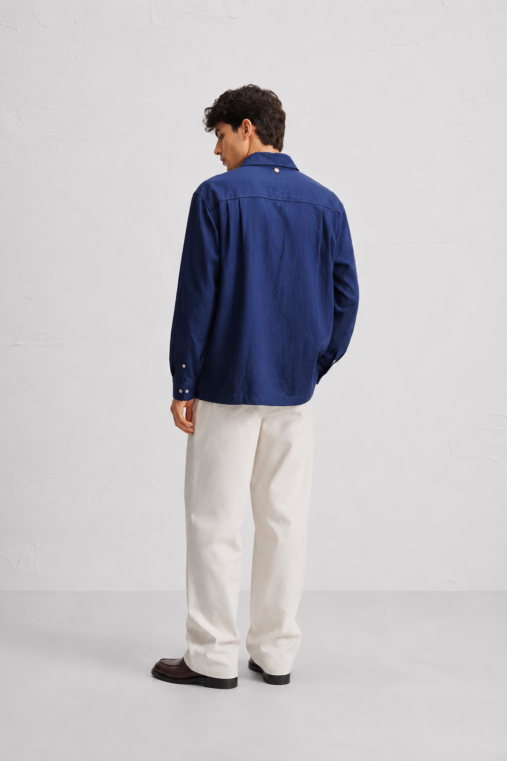 A man is shown from the back wearing a navy blue long-sleeve button-up shirt and off-white trousers, standing against a light gray studio background.