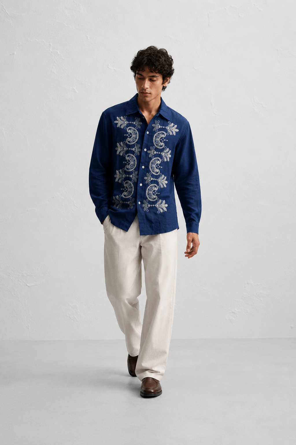 A man modeling a dark blue long-sleeve button-up shirt with white embroidered paisley and floral patterns, paired with off-white trousers and brown shoes, against a light gray textured studio background.